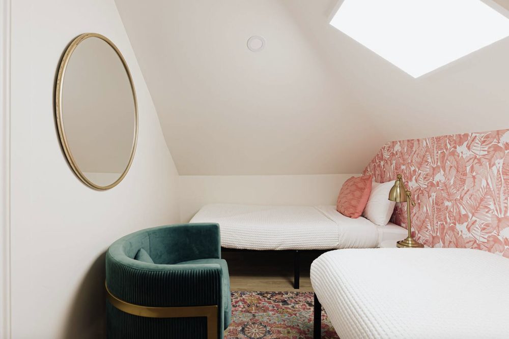 A room under the eaves with coral palm-print wallpaper, two beds, a teal velvet armchair and a skylight.