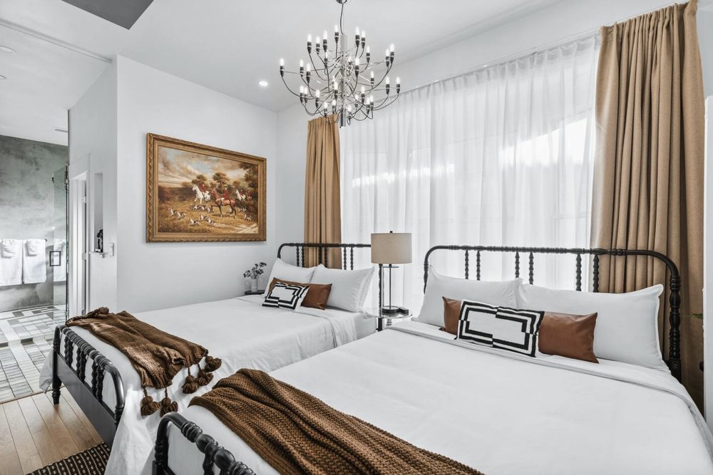 A white bedroom with two black spindle beds, a sputnik chandelier, an equestrian oil painting and an ensuite bathroom through the doorway.