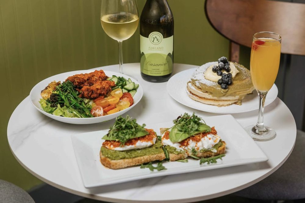 A table at Bedda: green walls, a bottle of Italian white, pancakes with blueberries, avocado toast and a mimosa.