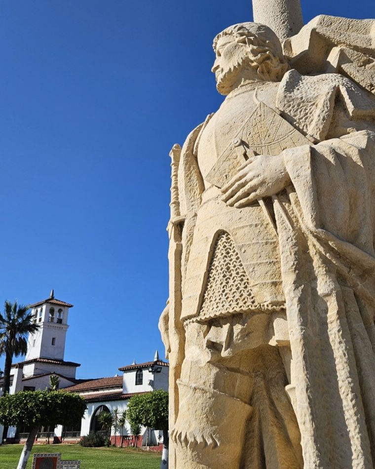 The Cabrillo statue in profile against a hard blue sky, with the Spanish-style visitor building behind.