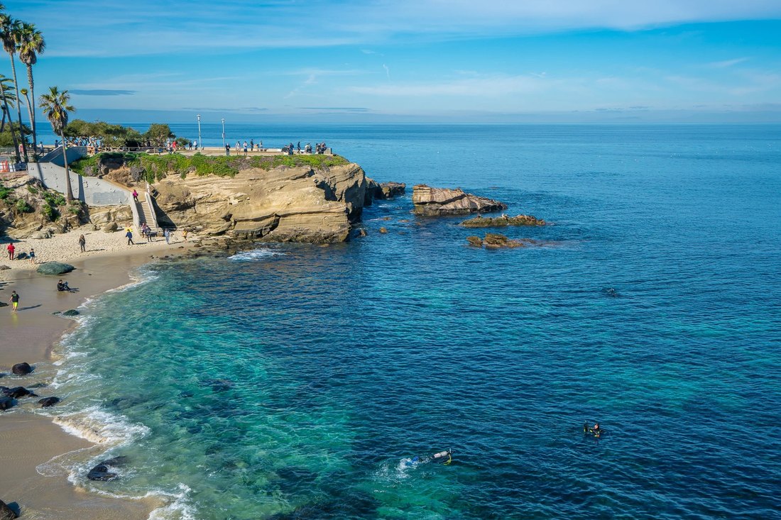 La Jolla Cove from the clifftop: a small sand beach, sandstone headland, and turquoise water with snorkellers and divers in it.