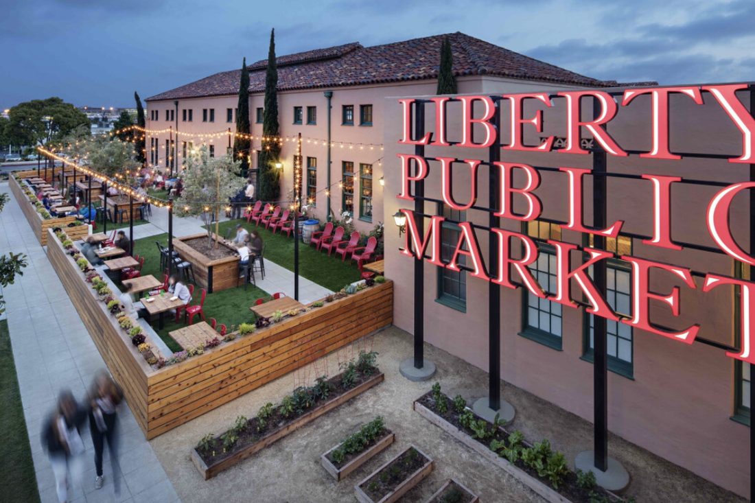 Liberty Public Market at dusk: a giant red neon sign on a pink stucco wall, string lights over the outdoor tables and raised garden beds below.