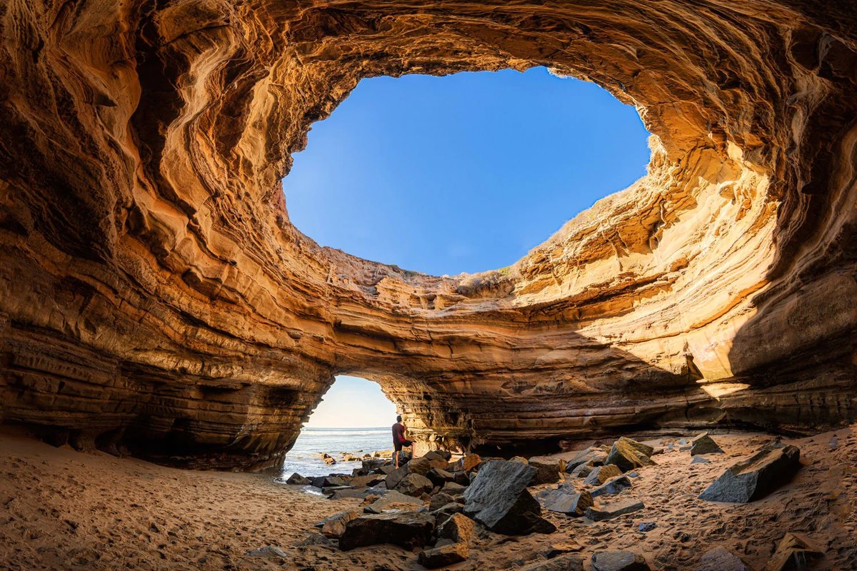 Inside a sandstone sea cave at Sunset Cliffs: a huge oval opening to blue sky overhead, and a smaller arch framing a person looking out to the ocean.